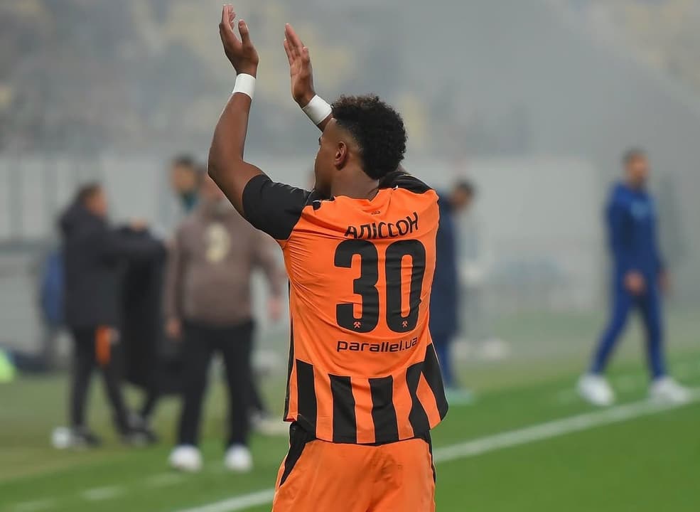 Alisson, jogador do Shakhtar Donetsk, com a camisa laranja e listras pretas, número 30, ergue as mãos em celebração.Shak