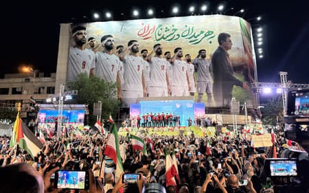 Manifestação em apoio ao governo iranianio, com presença da seleção do Irã na Copa, na praça Enghelab, centro de Teerã. Na imagem, uma multidão se concentra à noite empunhando bandeiras do país e celulares em frente a um grande palco iluminado, sob um imenso painel vertical que exibe jogadores da seleção nacional de futebol perfilados. (Foto: Patrícia Campos Mello/Folhapress)