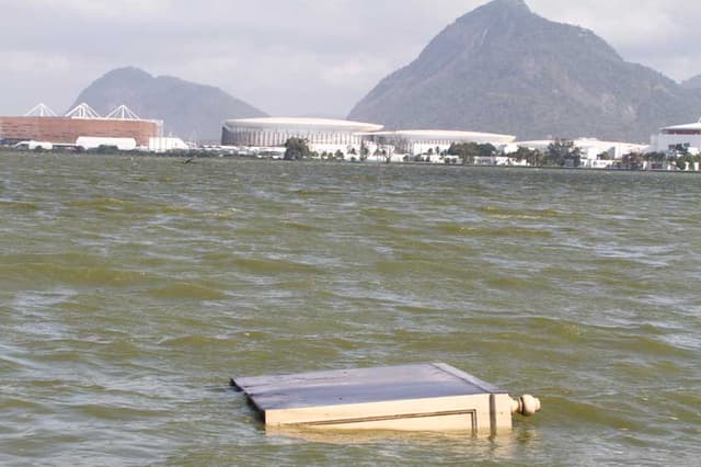 Móvel descartado flutua na Lagoa Jacarepaguá. Ao fundo, a Arena do Futuro e as Arenas Cariocas 1, 2 e 3, no Parque Olímpico