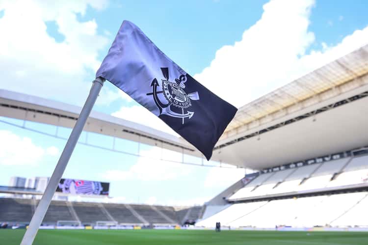 Bandeira do Corinthians tremulando sobre o campo, estádio vazio e arquibancadas ao fundo.