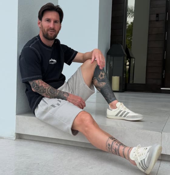 Messi sentado no chão, com uma perna flexionada e outra esticada, exibe tatuagens enquanto veste camisa preta, bermuda de moletom bege e tênis branco com detalhes cinza.