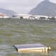 Móvel descartado flutua na Lagoa Jacarepaguá. Ao fundo, a Arena do Futuro e as Arenas Cariocas 1, 2 e 3, no Parque Olímpico