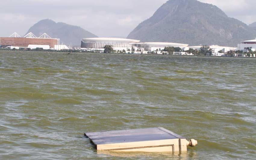 Móvel descartado flutua na Lagoa Jacarepaguá. Ao fundo, a Arena do Futuro e as Arenas Cariocas 1, 2 e 3, no Parque Olímpico