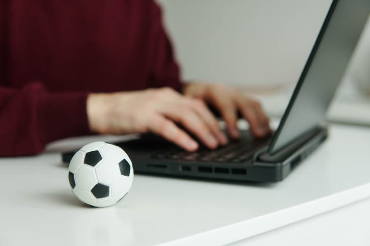 Young man reading soccer news or writing football article on laptop. Soccer ball on the table. Betting, gambling concept.