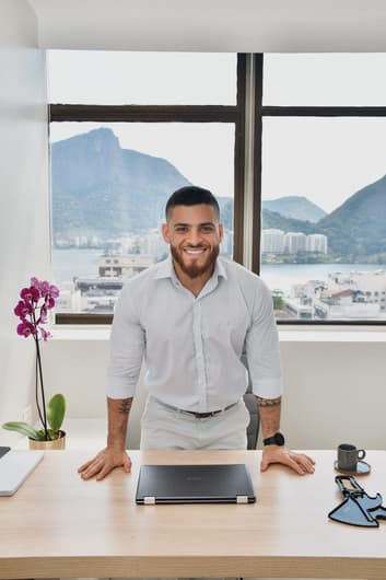 Nutricionista homem em pé em frente à janela de vidro com vista para o Rio de Janeiro, posando para a foto apoiado em uma mesa de madeira.