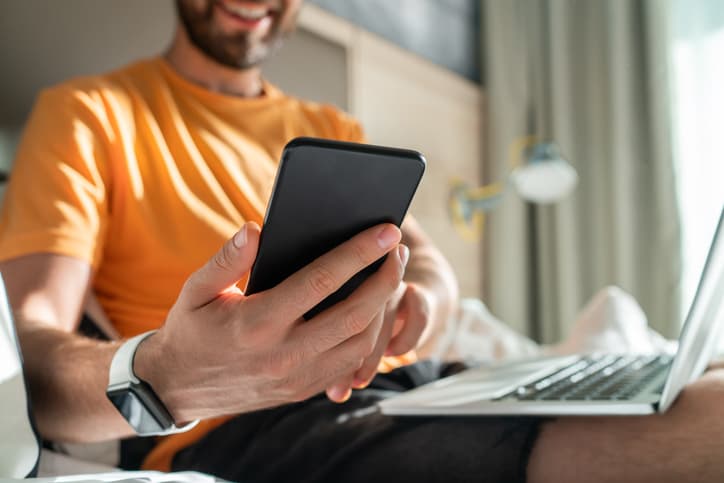 Close up cropped shot of smiling casually dressed business man spending his weekend at the hotel checking messages on his smartphone while lying in the bed