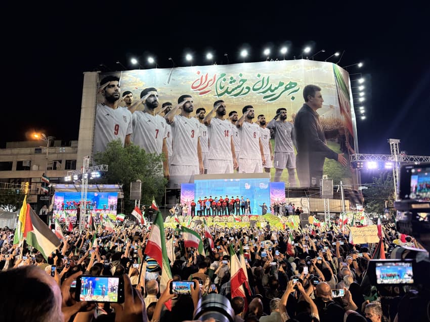 Manifestação em apoio ao governo iranianio, com presença da seleção do Irã na Copa, na praça Enghelab, centro de Teerã. Na imagem, uma multidão se concentra à noite empunhando bandeiras do país e celulares em frente a um grande palco iluminado, sob um imenso painel vertical que exibe jogadores da seleção nacional de futebol perfilados. (Foto: Patrícia Campos Mello/Folhapress)