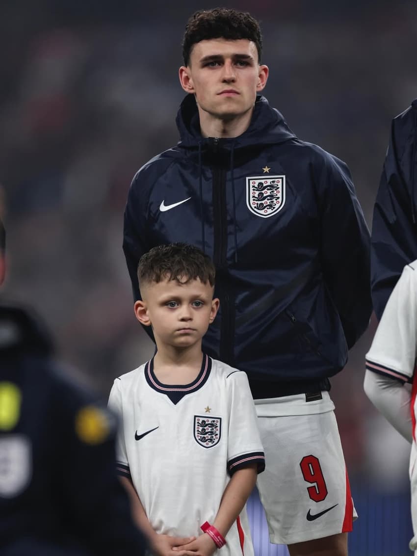 Phil Foden utilizando um casaco da Inglaterra com os braços para atrás e com o seu filho, Ronnie Foden, em sua frente