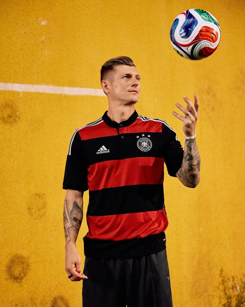 Toni Kroos utilizando a camisa da Alemanha, que tem listras horizontais pretas e vermelhas, e jogando uma bola de futebol pra cima