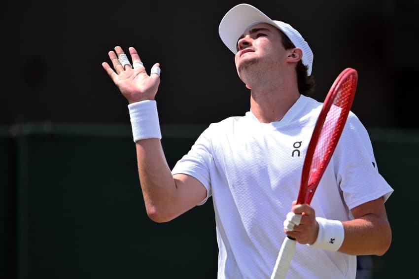 De camisa e munhequeiras brancas, segurando uma raquete com a mão esquerda, João Fonseca gesticula na partida contra o russo Roman Safiullin em Wimbledon