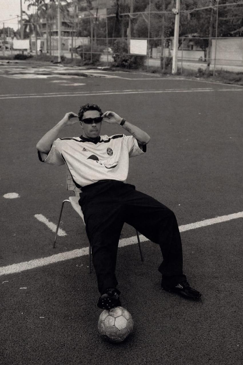 Foto preto e branca, Gabriel usa óculos de sol, camisa de futebol e calça preta e chuteira. Ele está sentado no meio do campo com bola no pé direito