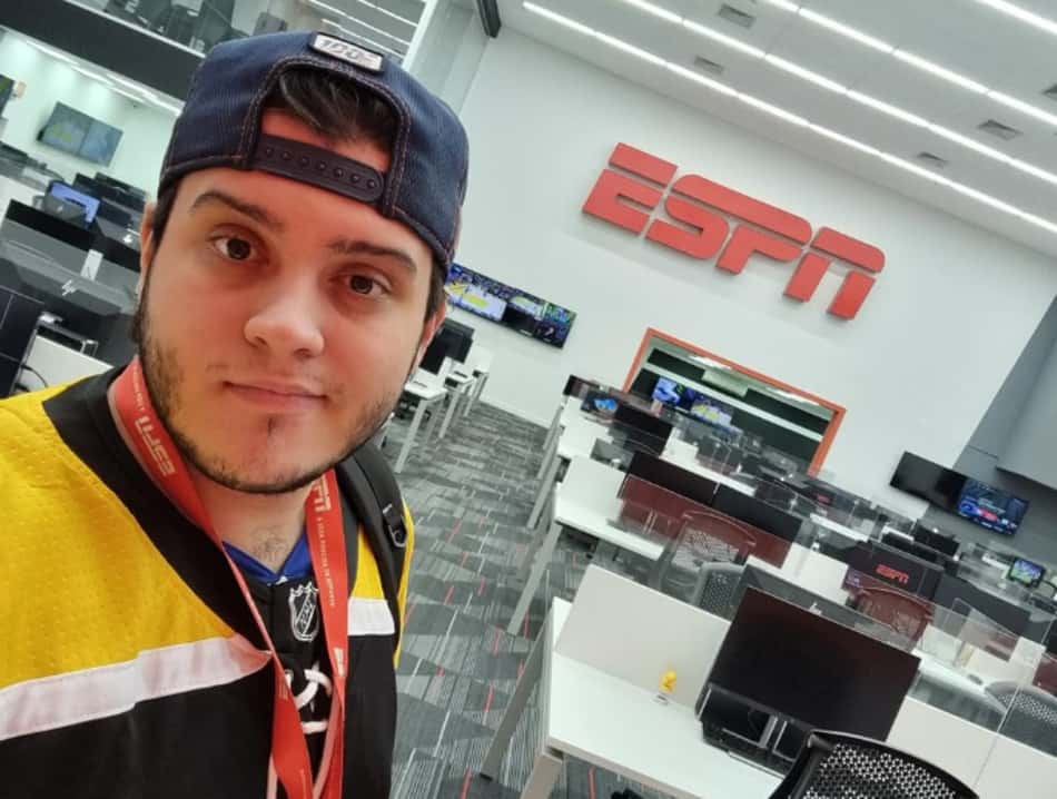 Matheus Pinheiro, usando boné azul virado para trás e camisa preta e amarela, tira uma selfie na redação da ESPN, com o logo da emissora ao fundo e mesas de trabalho com monitores