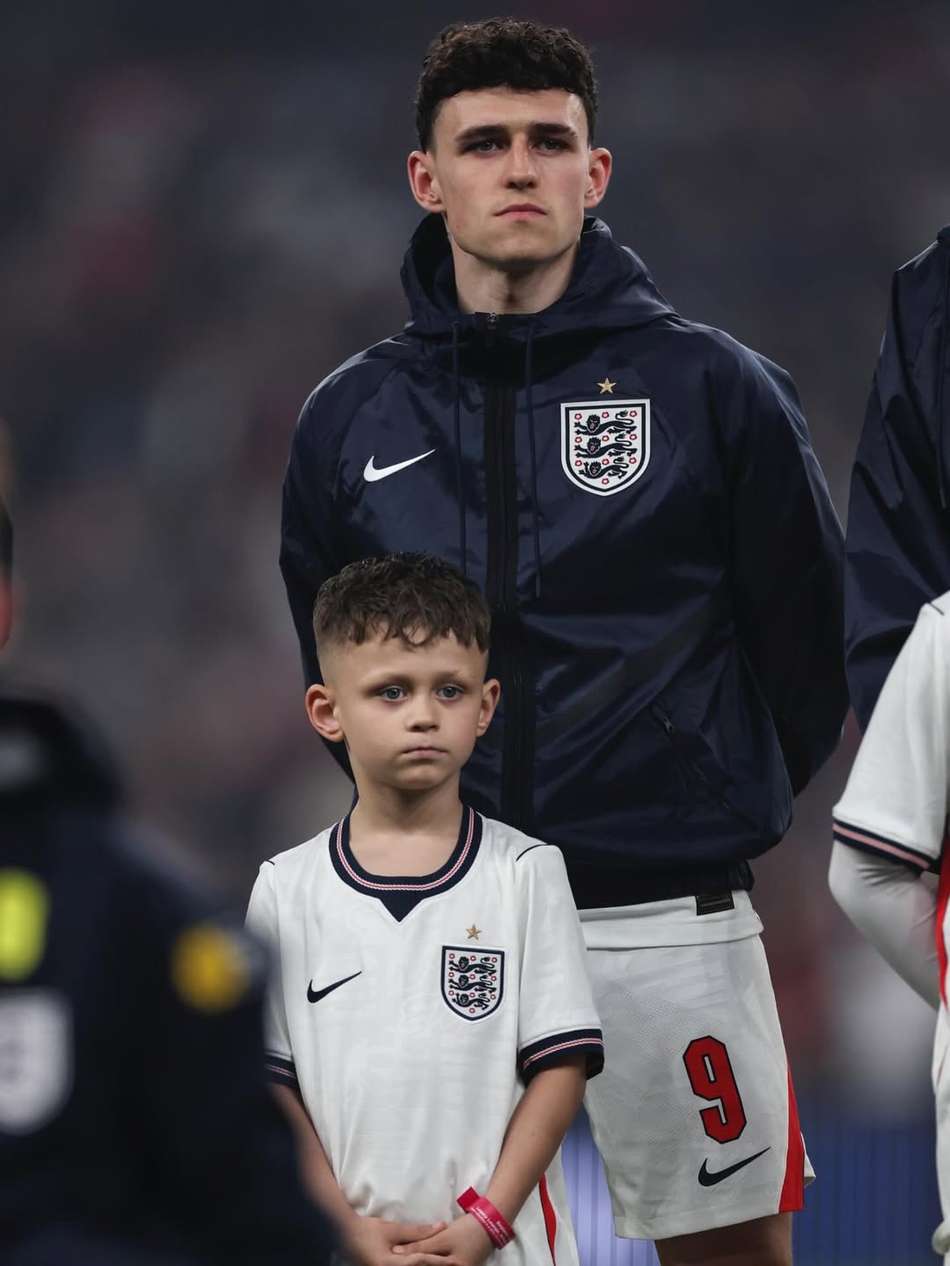 Phil Foden utilizando um casaco da Inglaterra com os braços para atrás e com o seu filho, Ronnie Foden, em sua frente