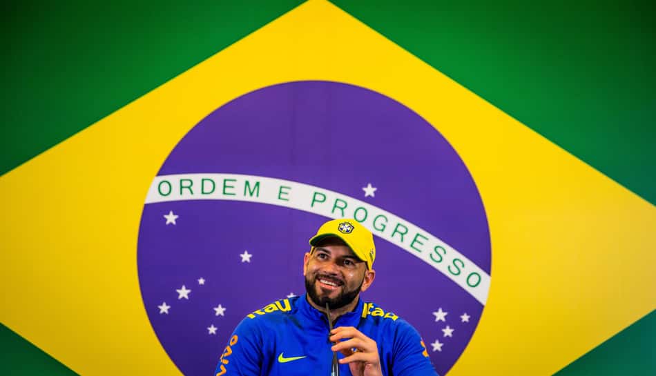 Weverton sorri segurando o microfone com a camisa de treino azul e o boné amarelo da Seleção Brasileira com uma bandeira do Brasil atrás