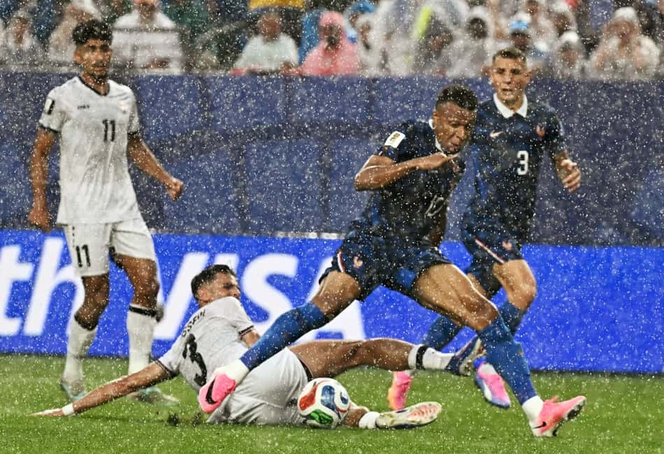 Mbappé com camisa azul da França divide bola com jogador do Iraque de branco que dá carrinho no gramado verde sob chuva forte