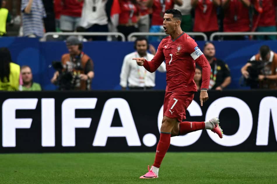 O atacante de Portugal, Cristiano Ronaldo, corre sorrindo de braços abertos para comemorar o gol de abertura de sua equipe contra o Uzbequistão. Ele está vestido com o uniforme principal da seleção portuguesa. Ao fundo, as arquibancadas do Houston Stadium estão repletas de torcedores.