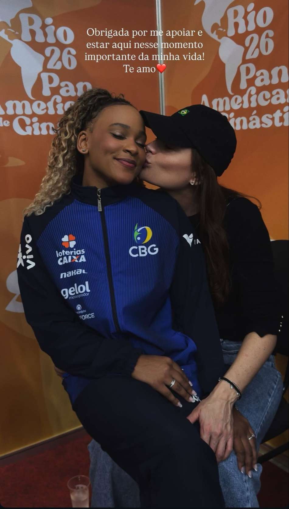 Rebeca Andrade sentada no colo da atleta Gisele recebendo um beijo no rosto