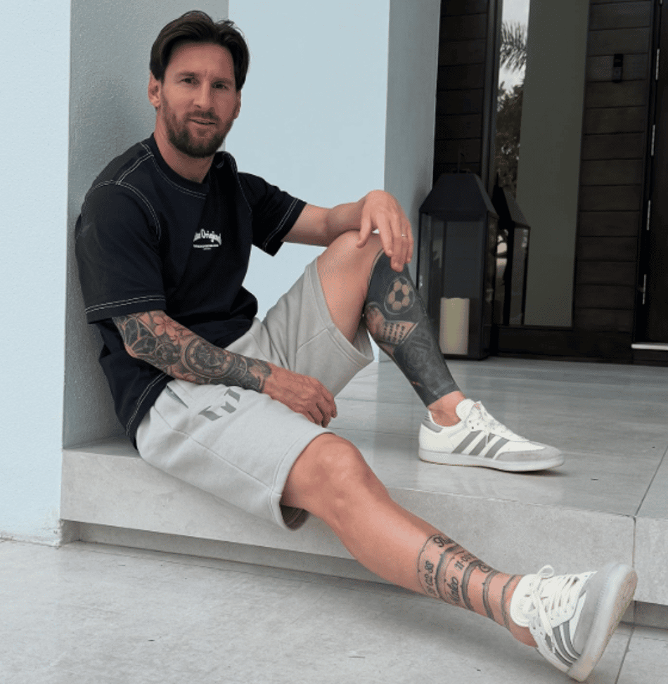 Messi sentado no chão, com uma perna flexionada e outra esticada, exibe tatuagens enquanto veste camisa preta, bermuda de moletom bege e tênis branco com detalhes cinza.