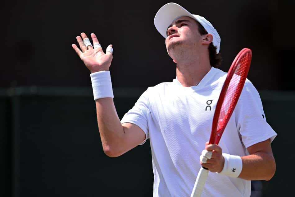 De camisa e munhequeiras brancas, segurando uma raquete com a mão esquerda, João Fonseca gesticula na partida contra o russo Roman Safiullin em Wimbledon