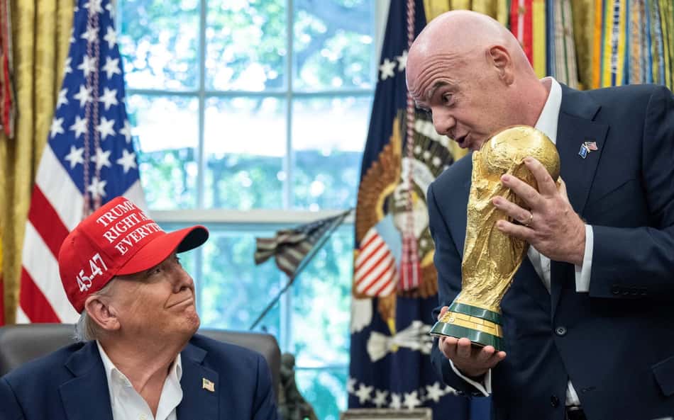 Gianni Infantino segurando a taça da Copa do Mundo de frente para Donald Trump, que usa um boné vermelho, e bandeiras dos Estados Unidos ao fundo