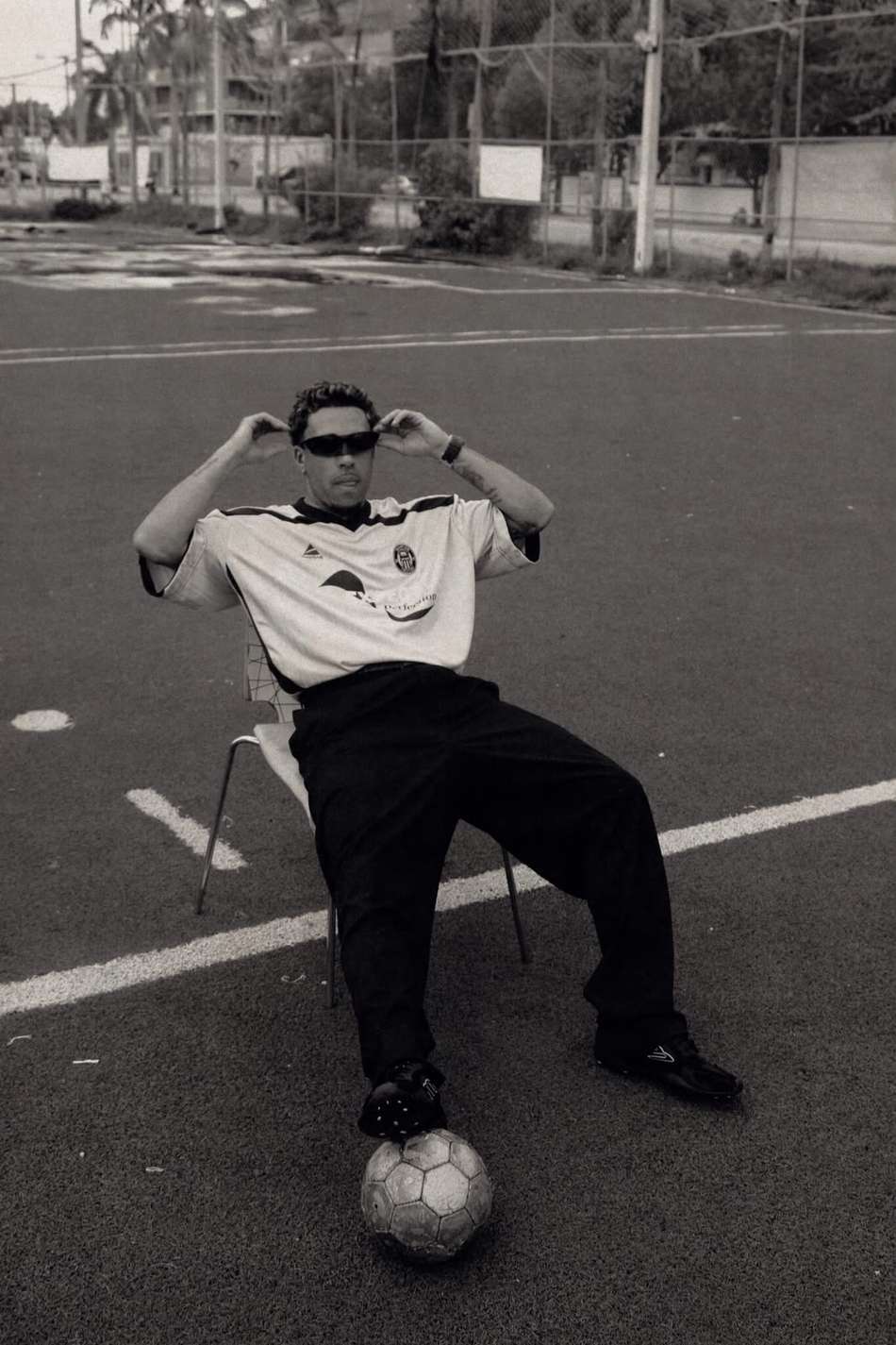 Foto preto e branca, Gabriel usa óculos de sol, camisa de futebol e calça preta e chuteira. Ele está sentado no meio do campo com bola no pé direito