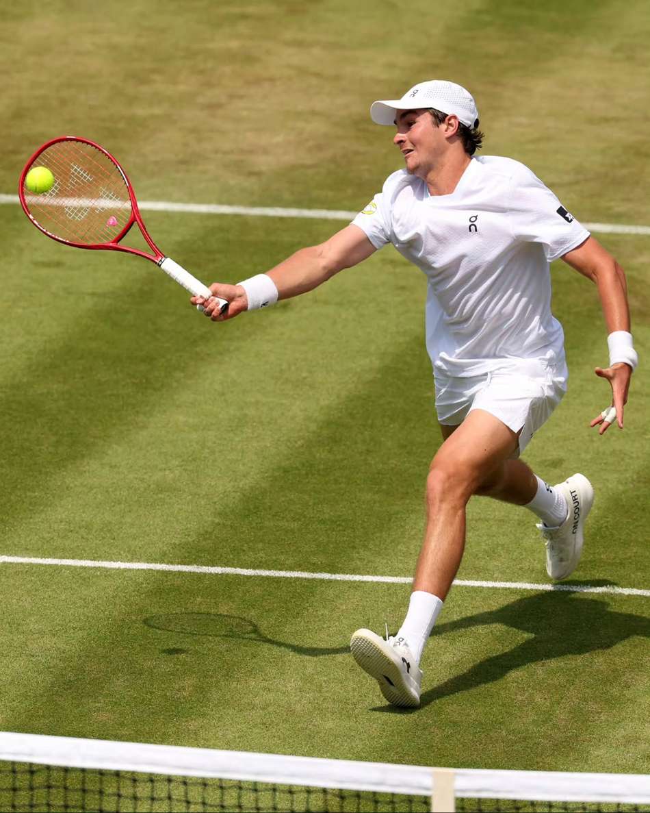 De camisa, boné, shorts, meias, tênis e munhequeiras brancos, João Fonseca aplica um forehand em jogo de Wimbledon