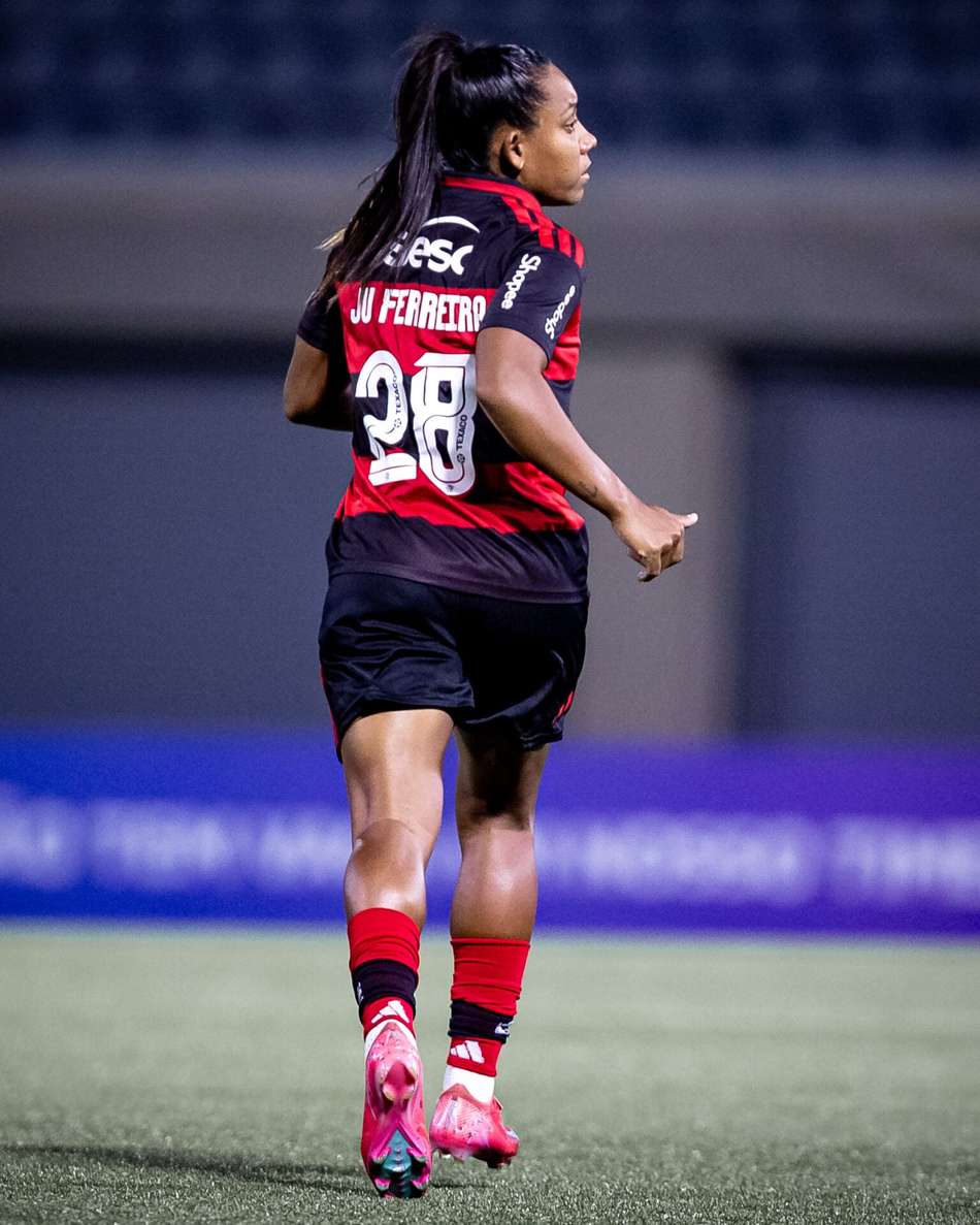 Ju Ferreira, volante do Flamengo, de costas correndo em campo de cabelo preso usando uniforme rubro-negro do clube: camisa vermelha e preta, short preto, meião vermelho com listras pretas e chuteira rosa