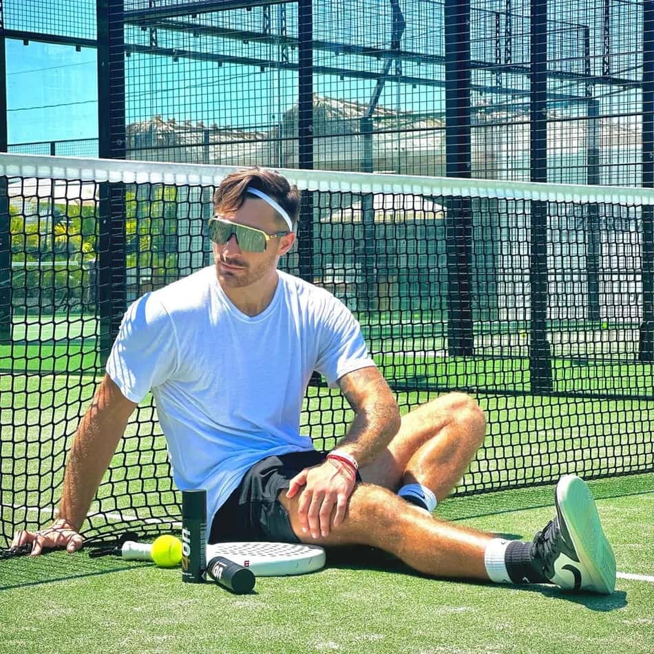 Nicolas Xiviller com óculos escuros sentado em frente à rede de tênis com raquete e bola ao lado