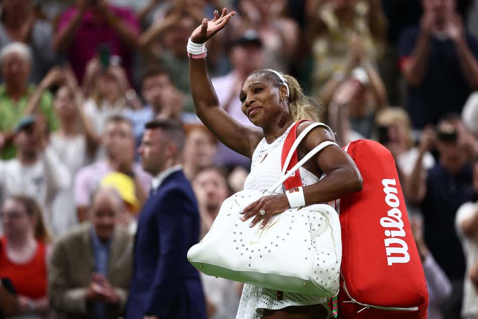 Segurando uma bolsa branca com a mão esquerda e carregando outra vermelha nas costas, Serena Williams acena para o público no All England Club com a mão direita