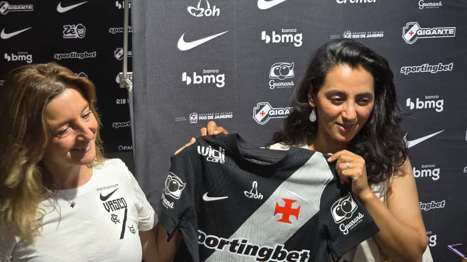 Khalida Popal na frente de um background com marcas parceiras do Vasco segurando uma camisa do clube