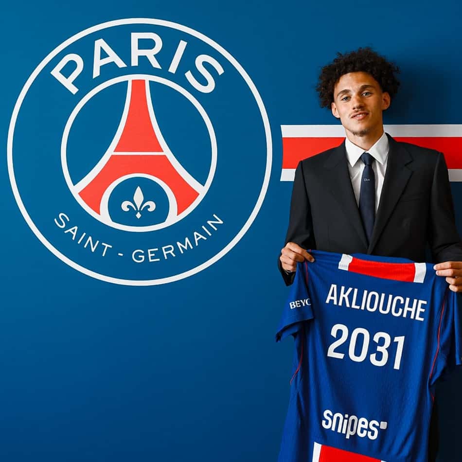 Maghnes Akliouche segurando camisa do PSG com 2031 escrito junto ao seu nome, ao lado de um escudo do clube
