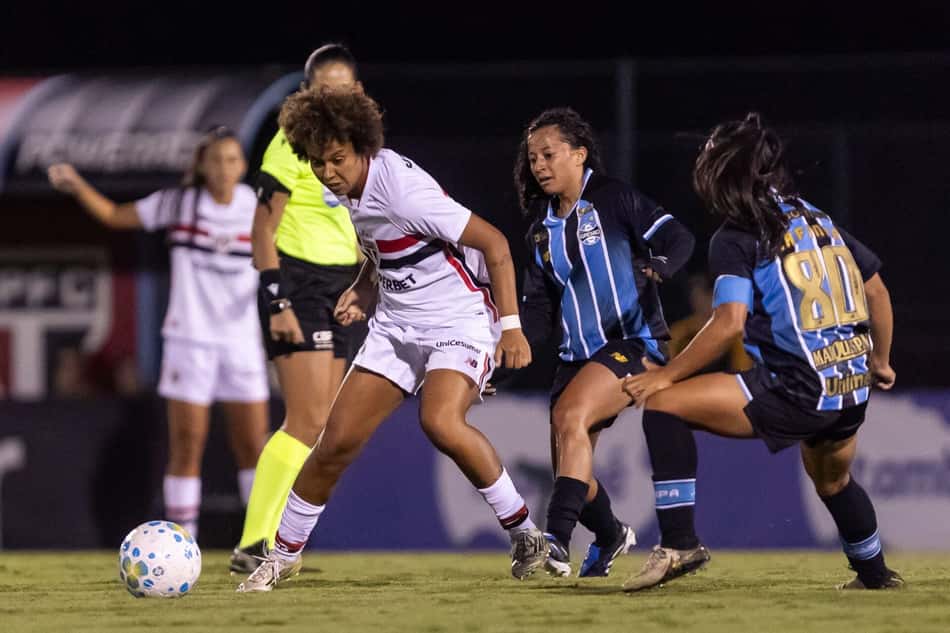 Jogadora do São Paulo protegendo a bola com duas marcadoras do Grêmio na cola e uma árbitra atrás na imagem.