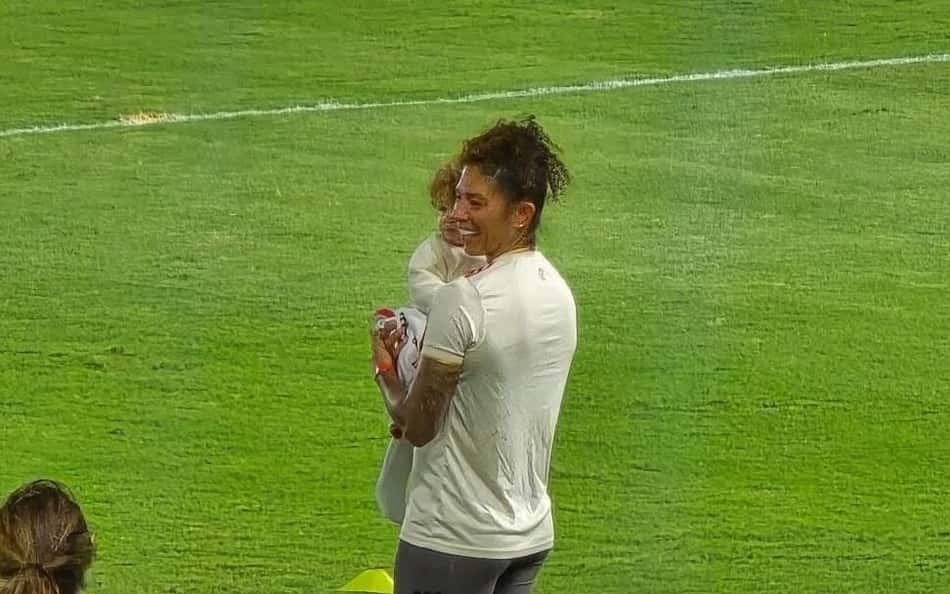 Cristiane, com camisa dourada do Flamengo, segura filha no colo no campo do estádio Luso-Brasileiro