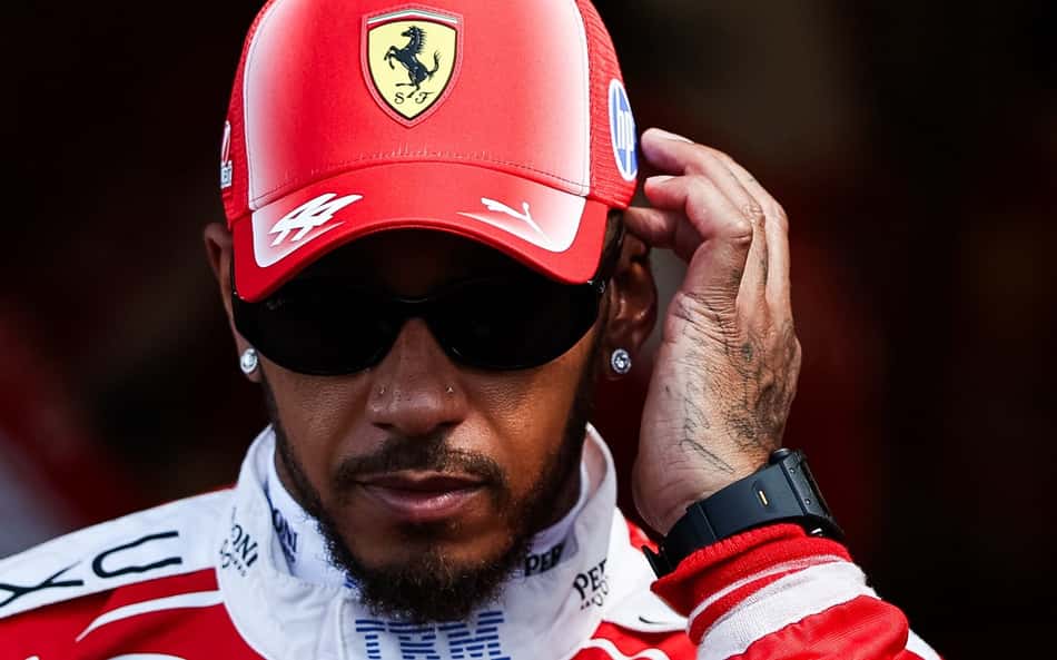 De boné da Ferrari e óculos escuros, Lewis Hamilton leva a mão esquerda ao ouvido