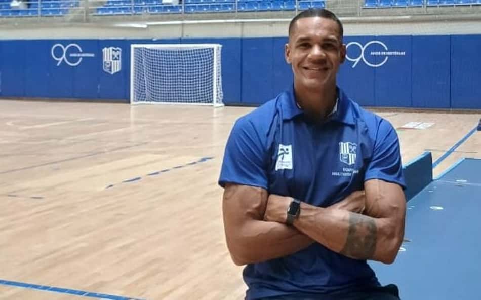 Samuel Fuchs, sentado em mesa e de braços cruzados, posa para foto na quadra principal do Minas Tênis Clube