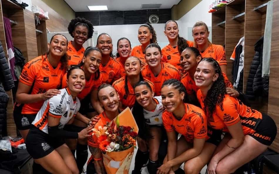 Tifanny posa com flores e o time do Osasco no vestiário da eqipe; todas sorriem