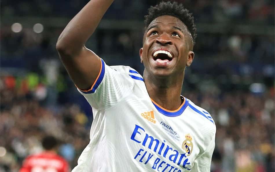 Vini Jr com camisa branca do Real Madrid aponta para o céu