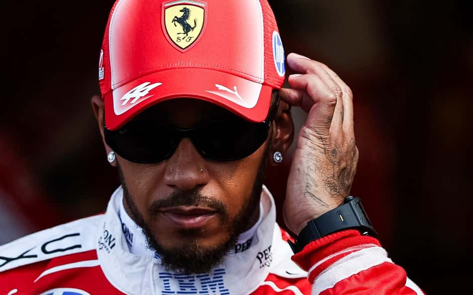 De boné da Ferrari e óculos escuros, Lewis Hamilton leva a mão esquerda ao ouvido