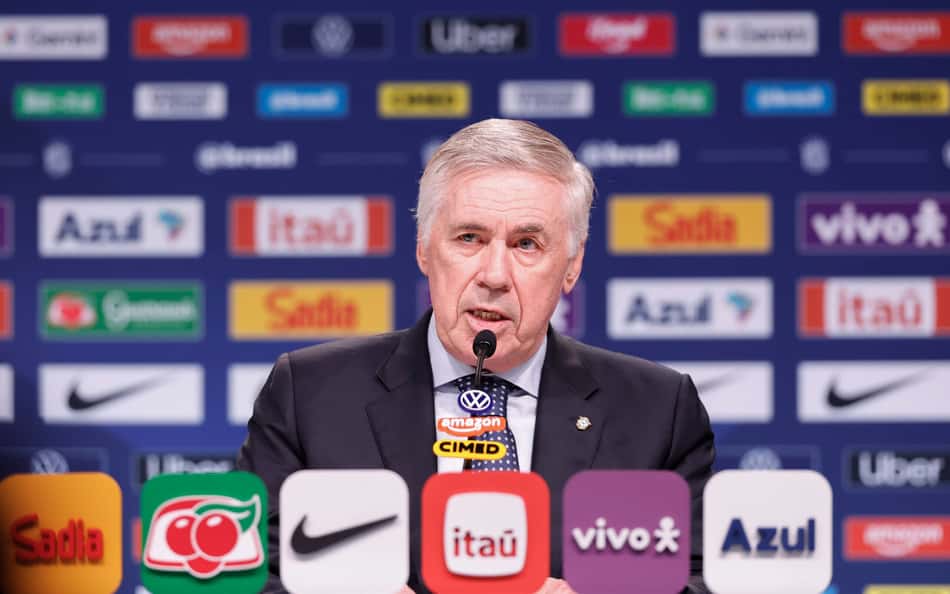 Homem de terno em coletiva de imprensa de convocação, com logotipos de patrocinadores ao fundo.