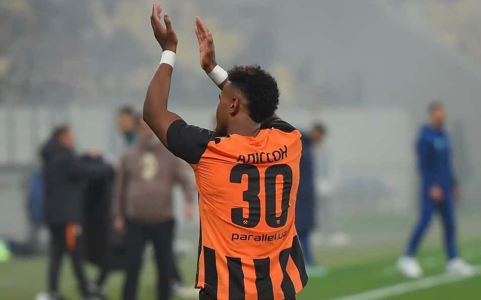 Alisson, jogador do Shakhtar Donetsk, com a camisa laranja e listras pretas, número 30, ergue as mãos em celebração.Shak