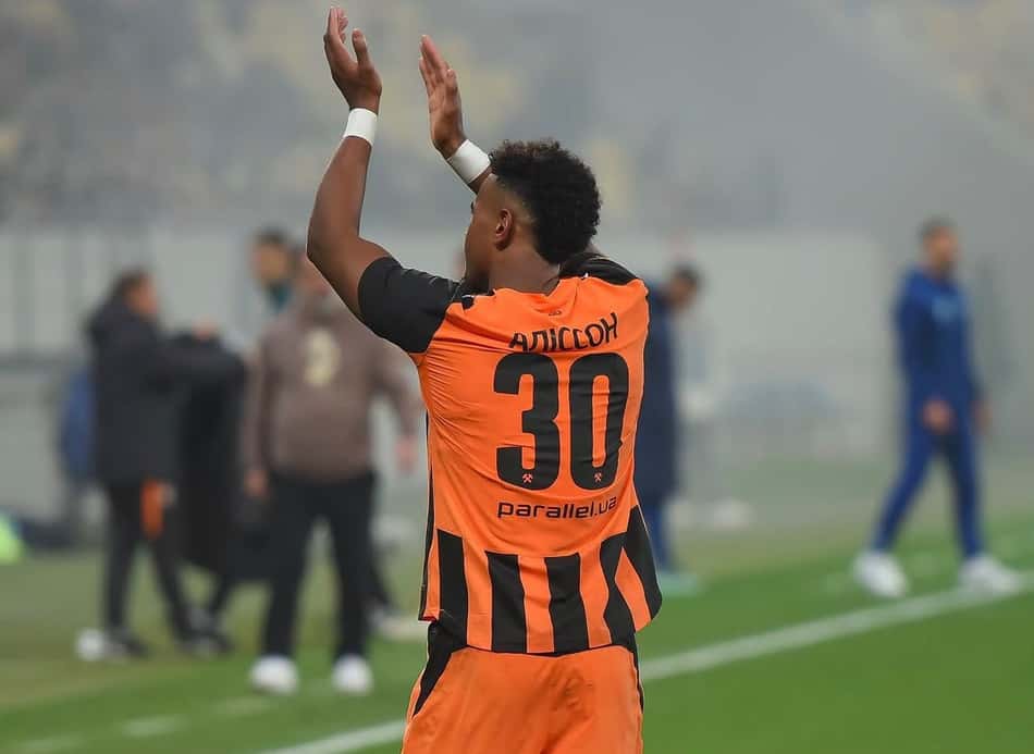 Alisson, jogador do Shakhtar Donetsk, com a camisa laranja e listras pretas, número 30, ergue as mãos em celebração.Shak