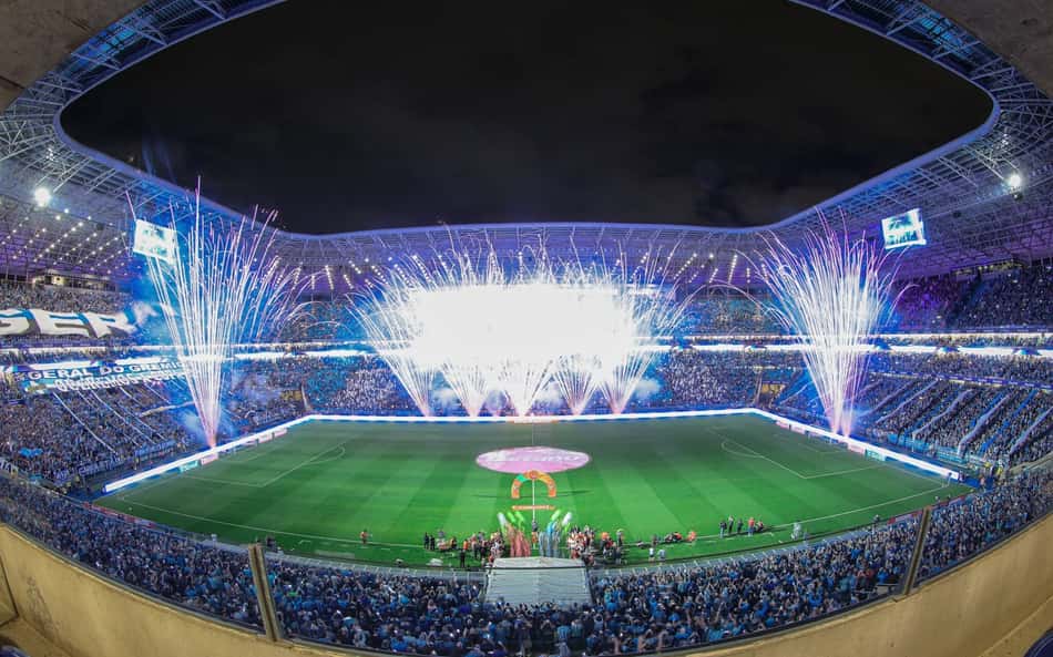 Arena Grêmio lotada, fogos de artifício iluminam o campo durante evento noturno.