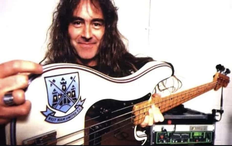 Baixista de cabelo comprido segura baixo branco com brasão azul, em estúdio com equipamento de áudio ao fundo.