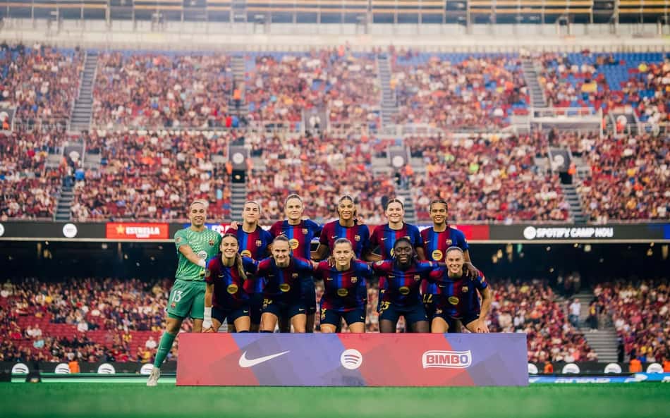 Barcelona Feminino posando para foto coletiva no estádio, torcida lotada ao fundo.