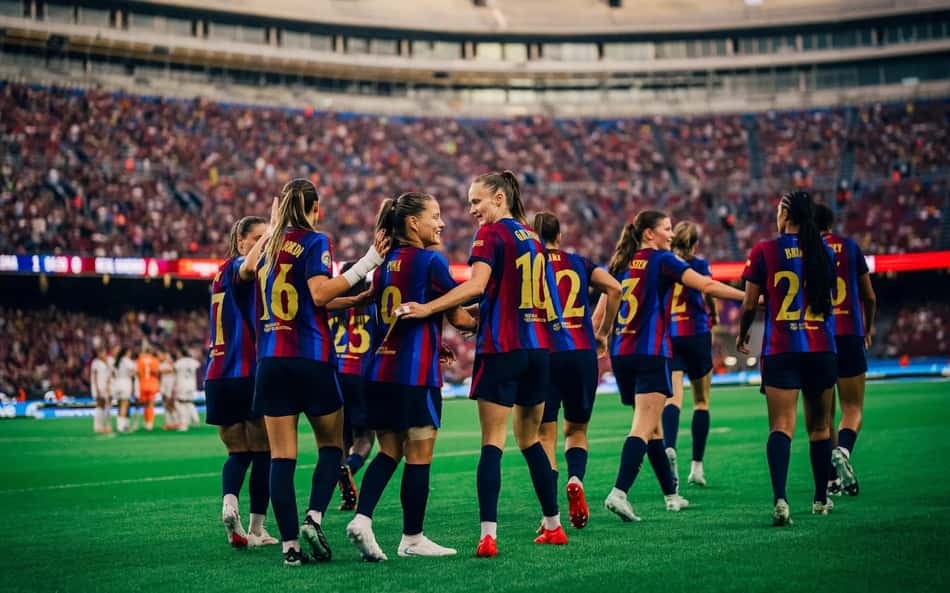 Jogadoras do Barcelona Feminino celebram no campo, estádio lotado ao fundo.