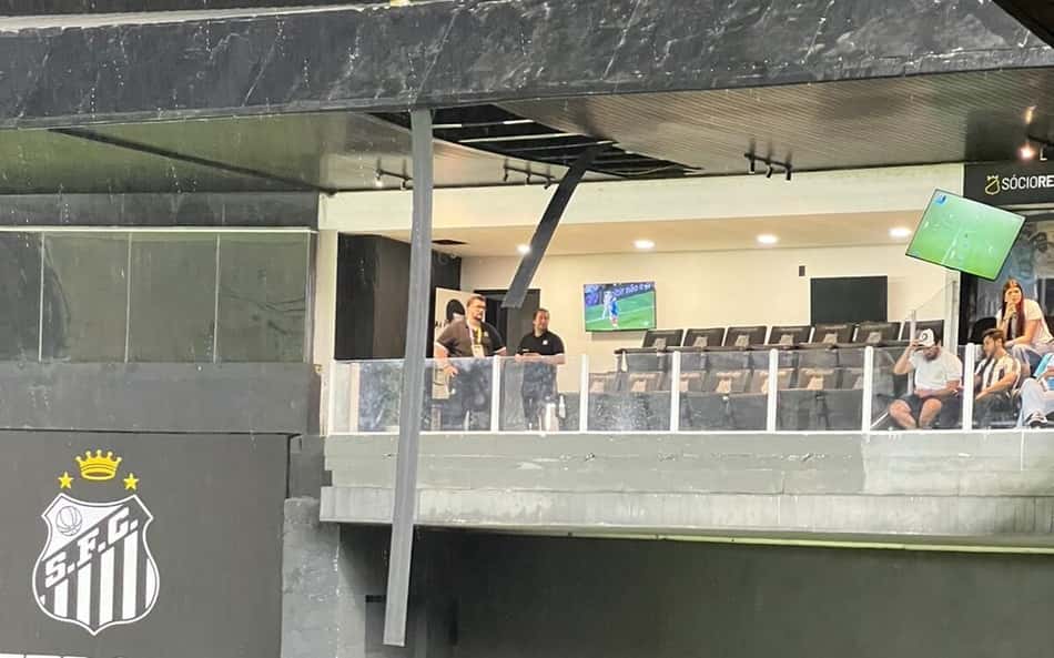 Torcedores na arquibancada do Campo Vila Belmiro assistem à partida, tela ao fundo e banner com o escudo do Santos FC.