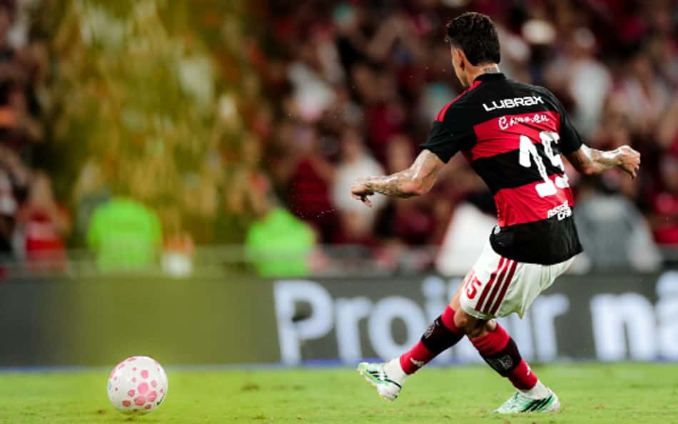 Jogador com uniforme listrado vermelho e preto, número 14, chutando uma bola rosa em campo, torcida ao fundo.