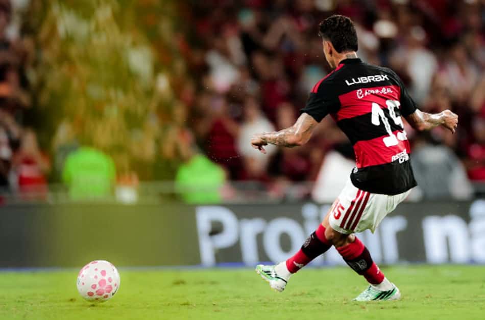 Jogador com uniforme listrado vermelho e preto, número 14, chutando uma bola rosa em campo, torcida ao fundo.
