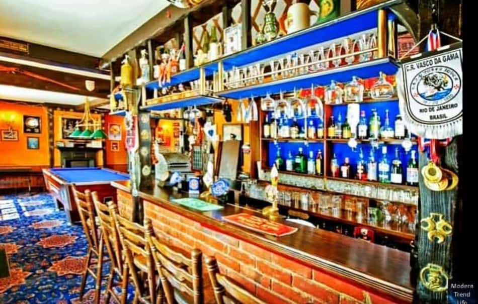 Bar com balcão de tijolo, mesas de sinuca e prateleiras com garrafas; bandeira do Vasco da Gama.