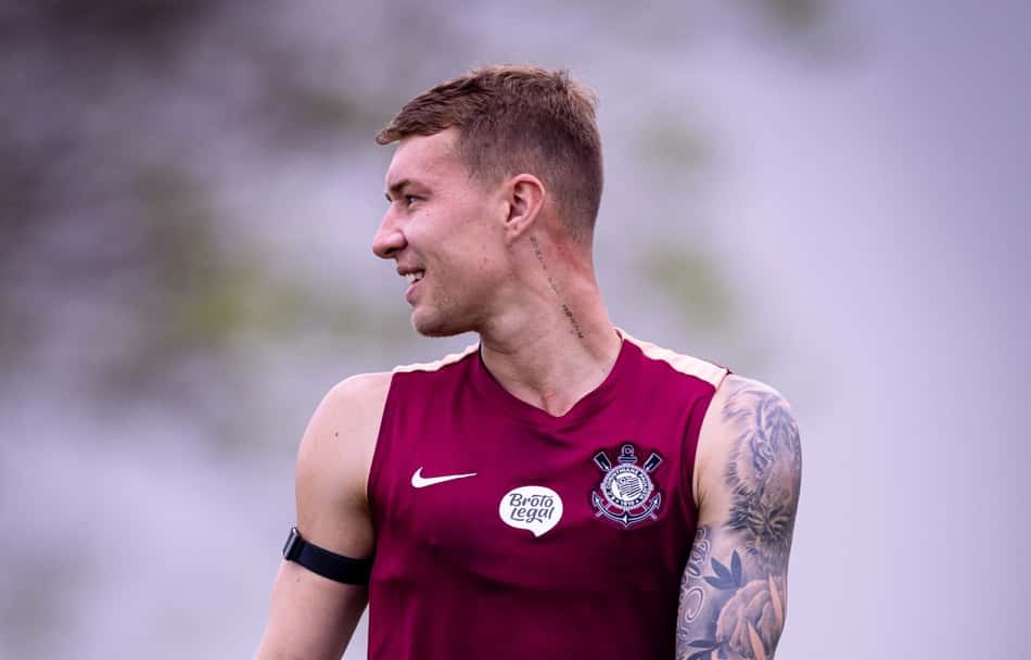 Charles com camisa de treino marrom do Corinthians, tatuado no braço, sorrindo e olhando para a esquerda.